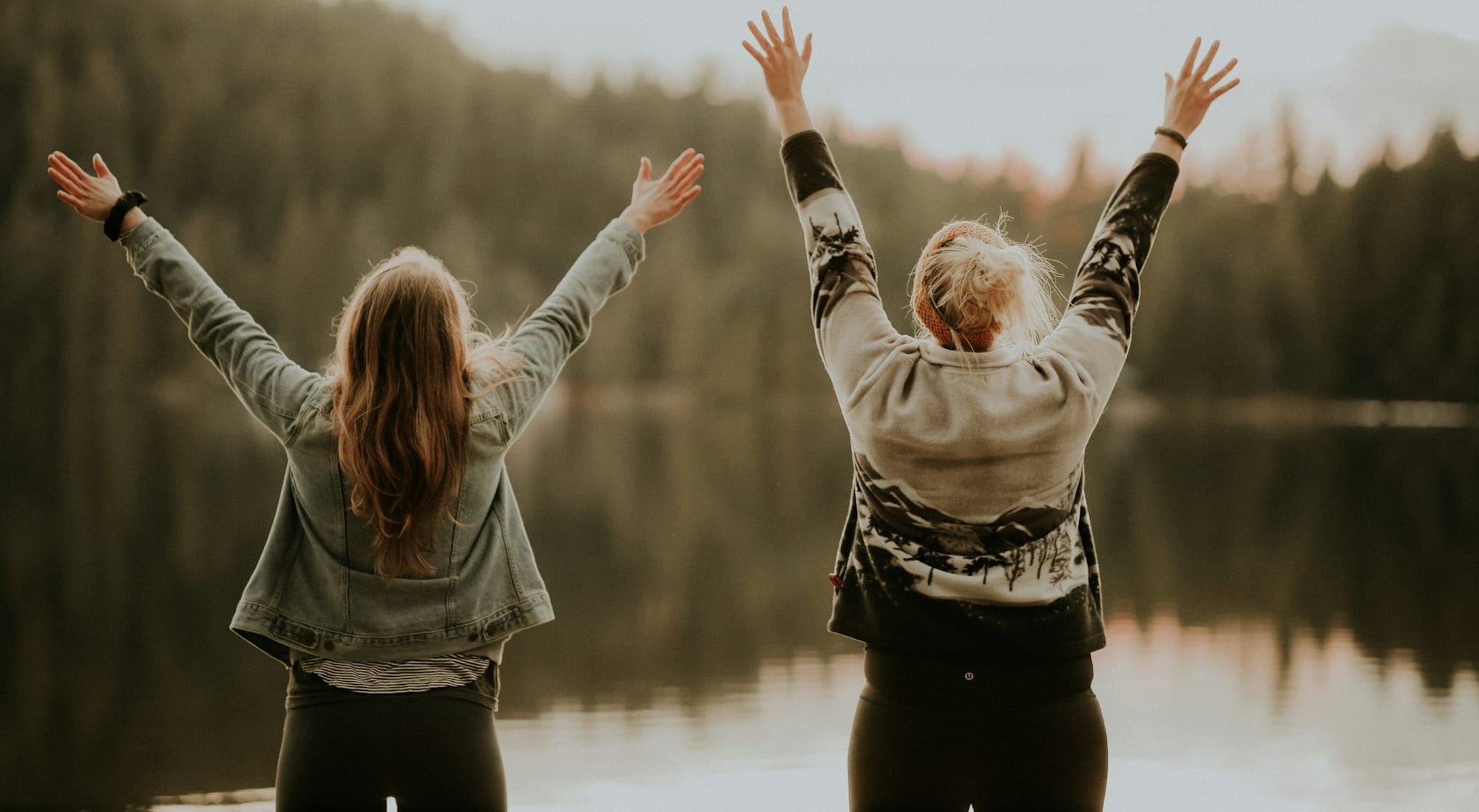 two women hands up standing beside body of water